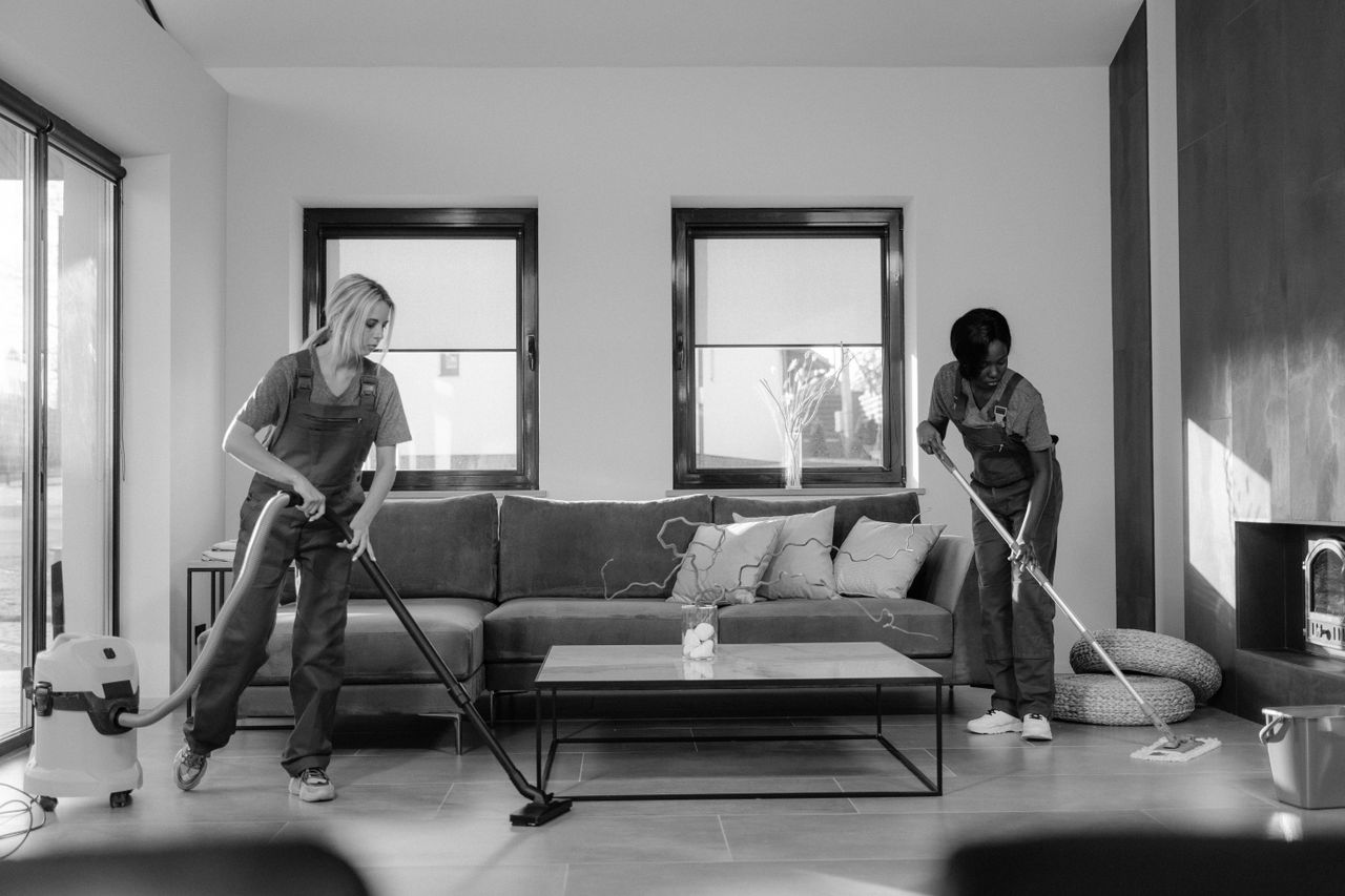 services-03 Two women in work clothes cleaning a modern living room using a vacuum and mop.
